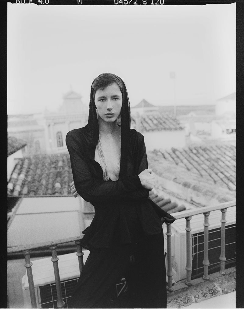Black-and-white portrait of a woman with arms crossed on a balcony, wearing a dark outfit with a hood, overlooking tiled rooftops.