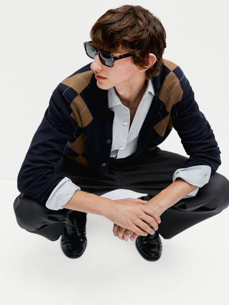 A young adult man in a crouching pose, wearing a cardigan sweater, grey pants, and black shoes against a clean, bright background.