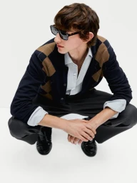 Man wearing sunglasses, argyle cardigan, light shirt, and dark trousers crouching against a white background for Massimo Dutti.