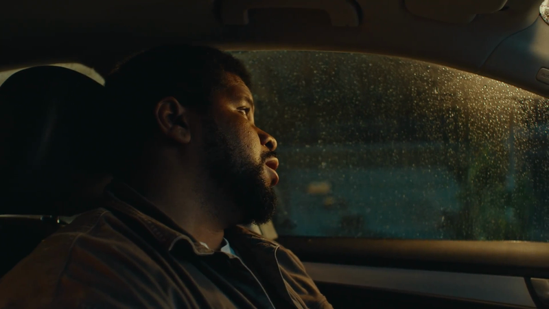 Profile view of an African-American man in a car, looking out the rain-streaked window in subdued lighting, creating a contemplative mood.