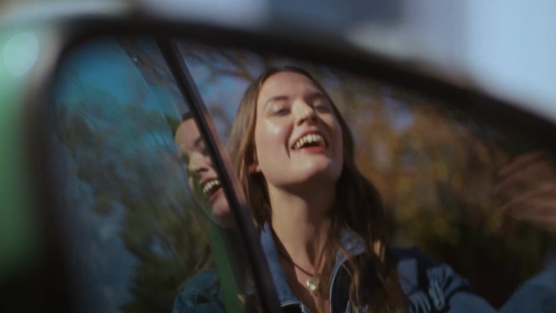 Smiling woman with long hair, wearing a denim jacket, photographed inside of a car in natural light with blurred background.