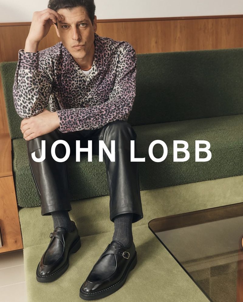 Man wearing a spotted shirt and leather pants sitting on a green sofa and wearing John Lobb leather shoes, with the brand name visible.