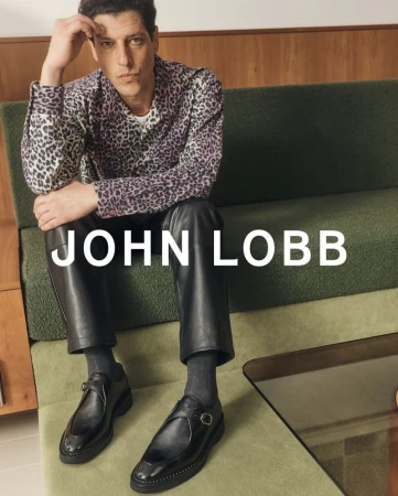 Man wearing a spotted shirt and leather pants sitting on a green sofa and wearing John Lobb leather shoes, with the brand name visible.