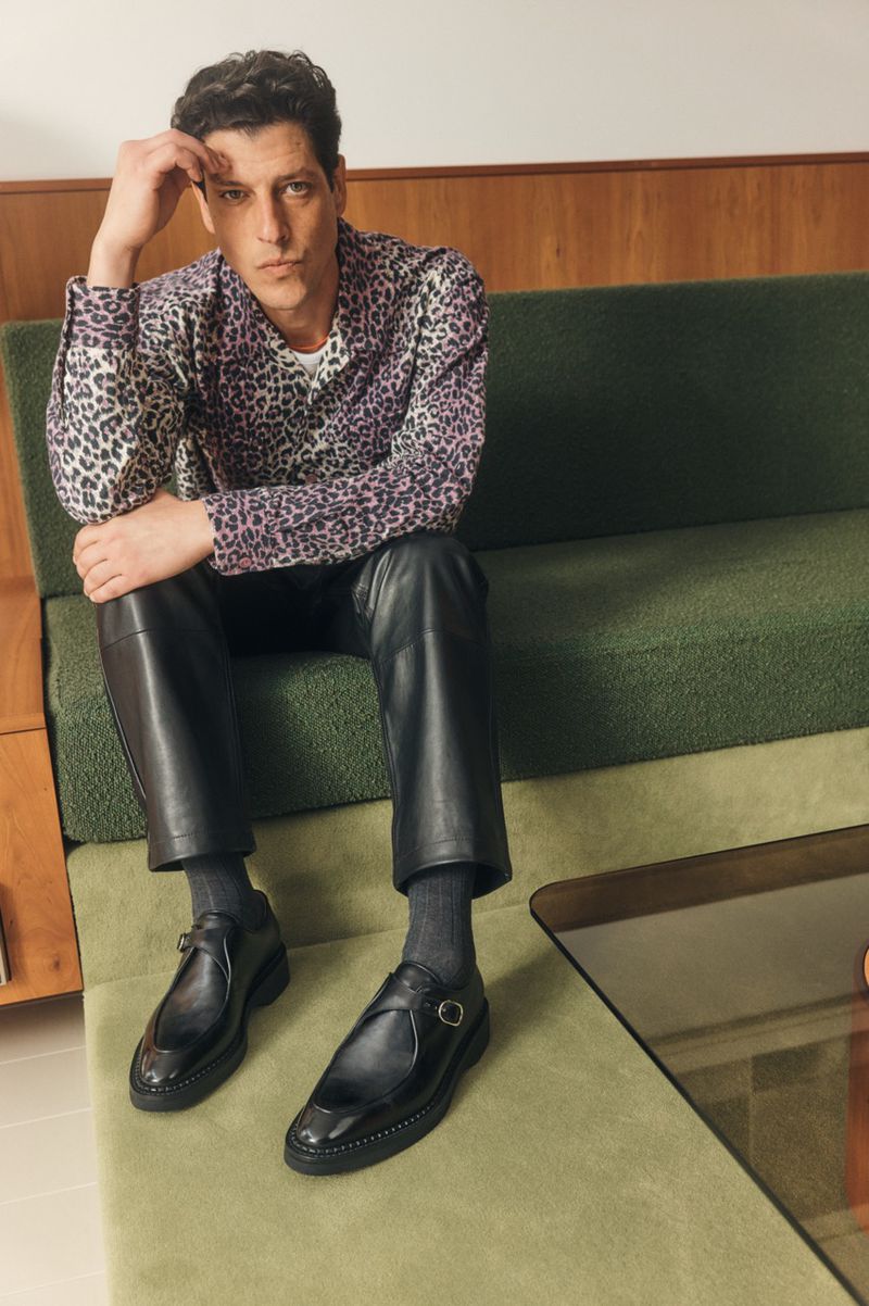 Man seated on a green sofa wearing a patterned shirt, black leather trousers, and black leather shoes.