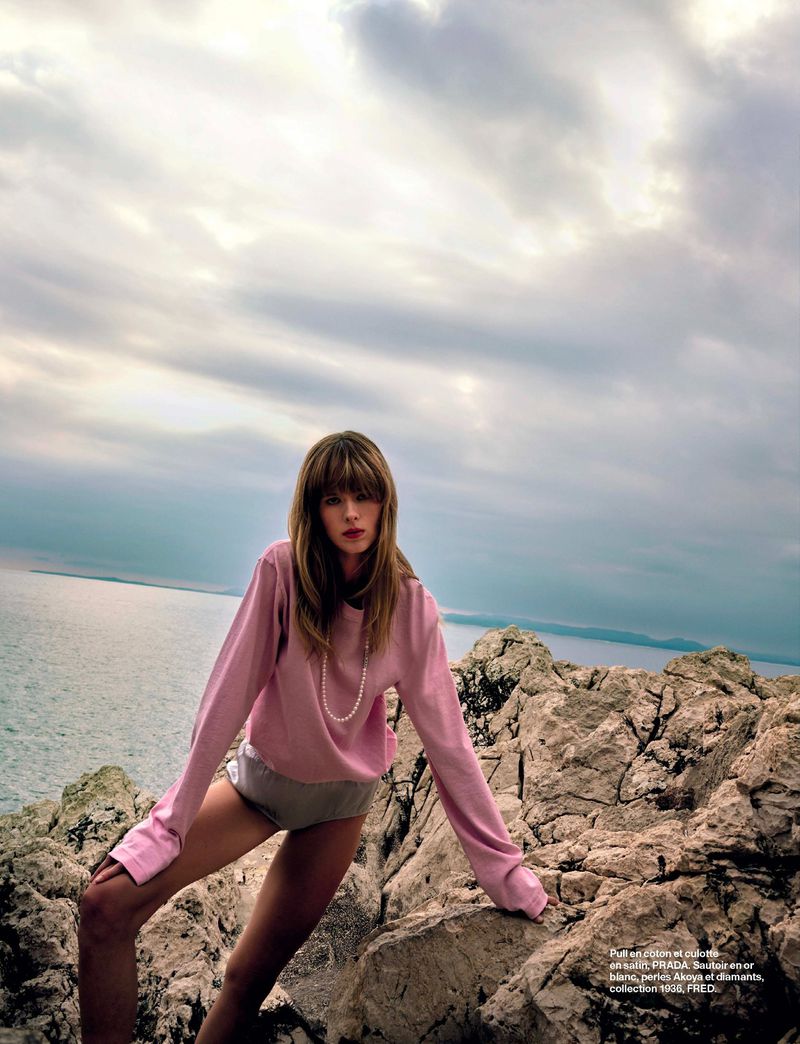 A woman poses on a rocky outcrop by the sea, wearing a light pink sweater and shorts, with a cloudy sky in the background.