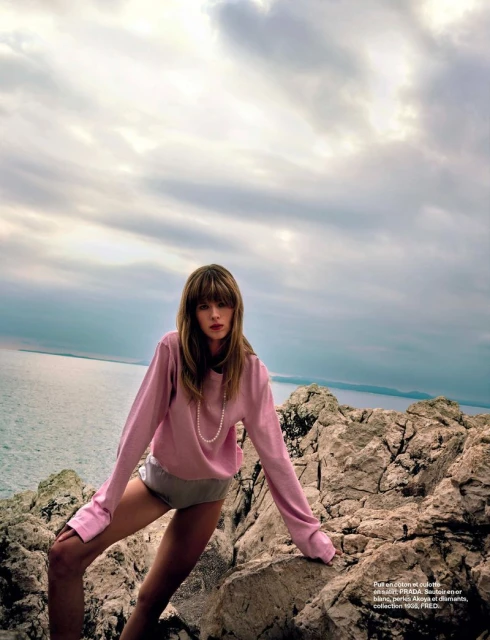 A woman poses on a rocky outcrop by the sea, wearing a light pink sweater and shorts, with a cloudy sky in the background.