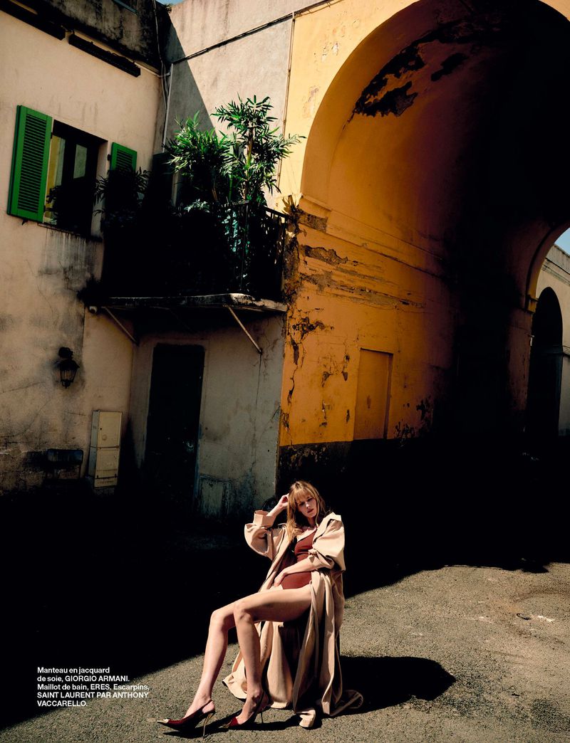 Fashion editorial shot of an adult woman in lingerie and a trench coat sitting in a city square with buildings and an archway in the background.
