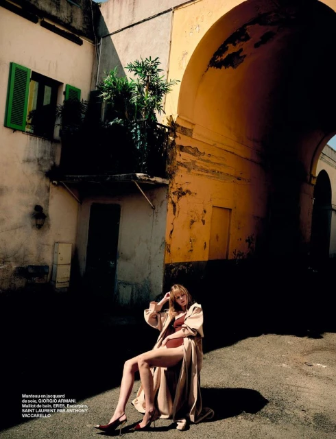 Fashion editorial shot of an adult woman in lingerie and a trench coat sitting in a city square with buildings and an archway in the background.