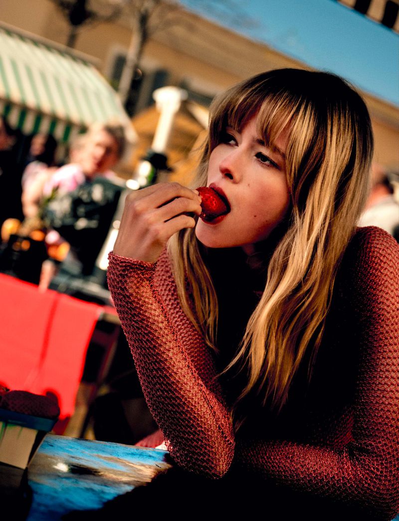 A woman with blonde hair eats a strawberry outdoors at a market, wearing a textured top, with blurred background people and striped awning.