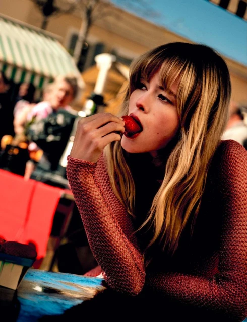 A woman with blonde hair eats a strawberry outdoors at a market, wearing a textured top, with blurred background people and striped awning.