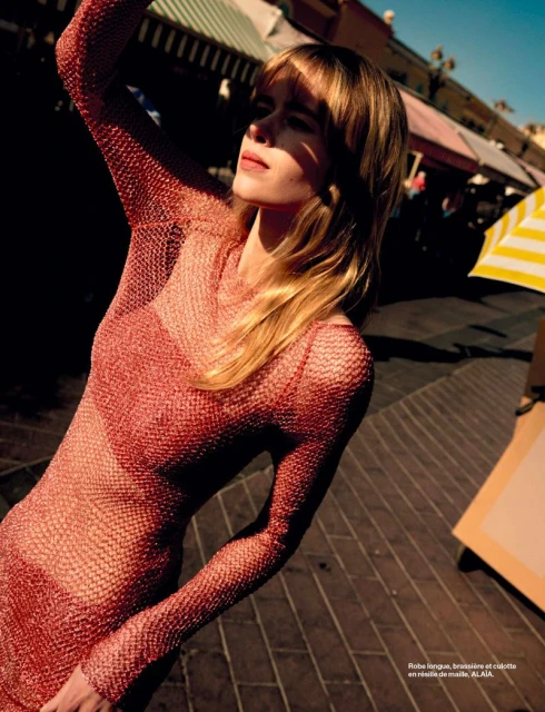 Blonde woman wearing a sheer rose-colored dress in an urban marketplace, capturing a sunlit fashion moment with a striped umbrella nearby.