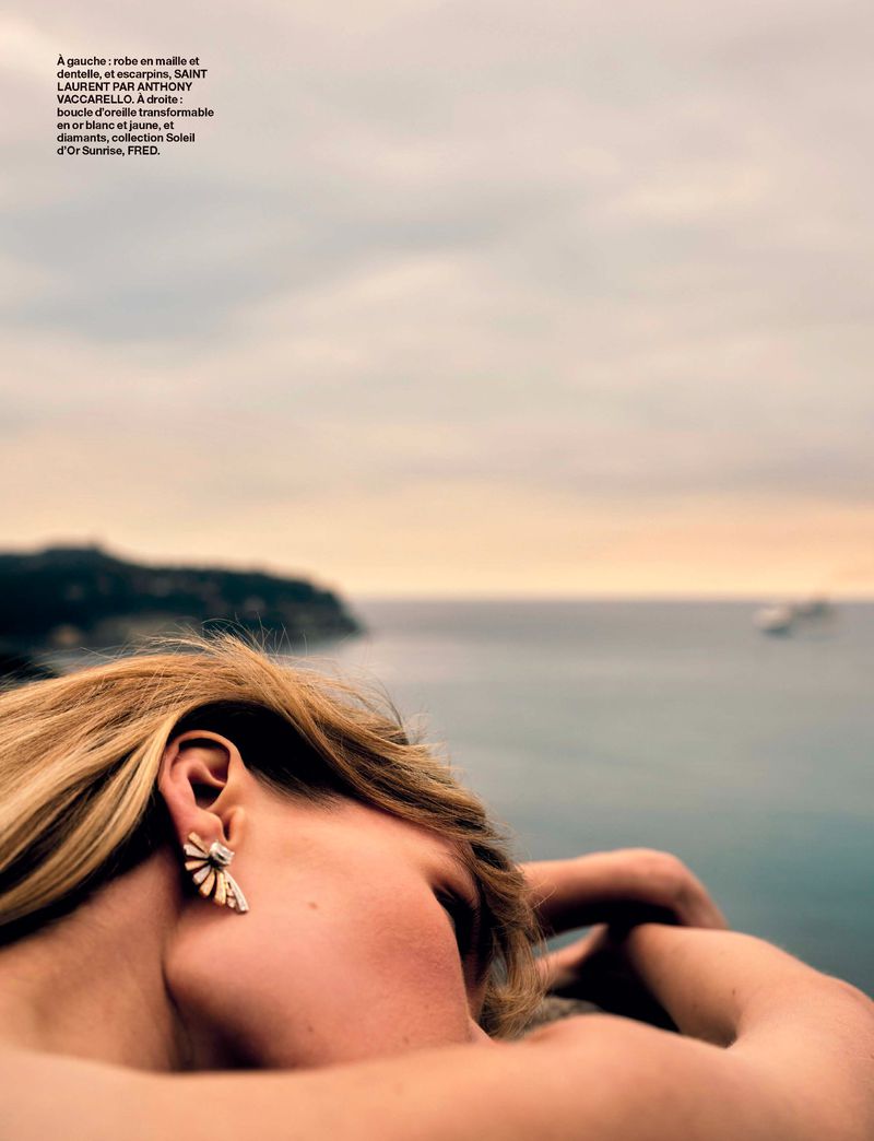 Woman with blonde hair and statement earrings gazes out at a seascape with islands under an overcast sky at golden hour. Fashion, beauty.