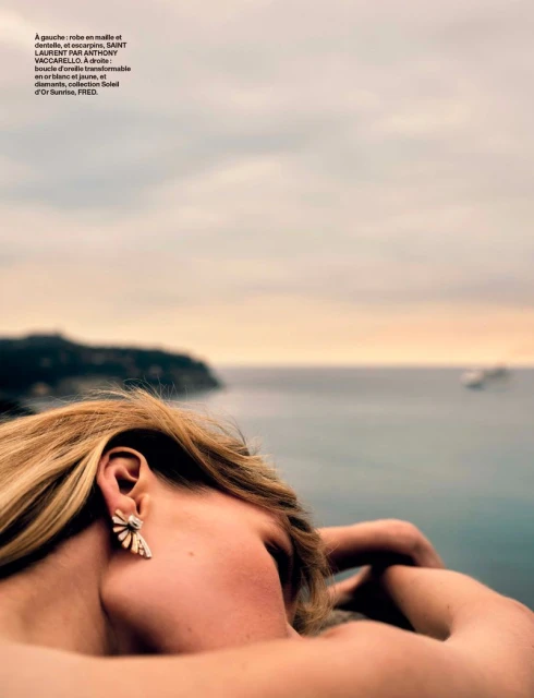 Woman with blonde hair and statement earrings gazes out at a seascape with islands under an overcast sky at golden hour. Fashion, beauty.