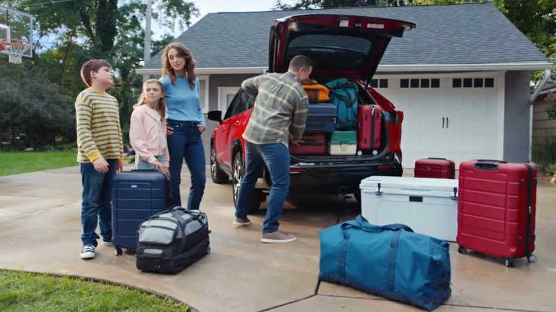 Family of four packing luggage into the trunk of a red SUV in front of a suburban house, preparing for a trip on a sunny day.