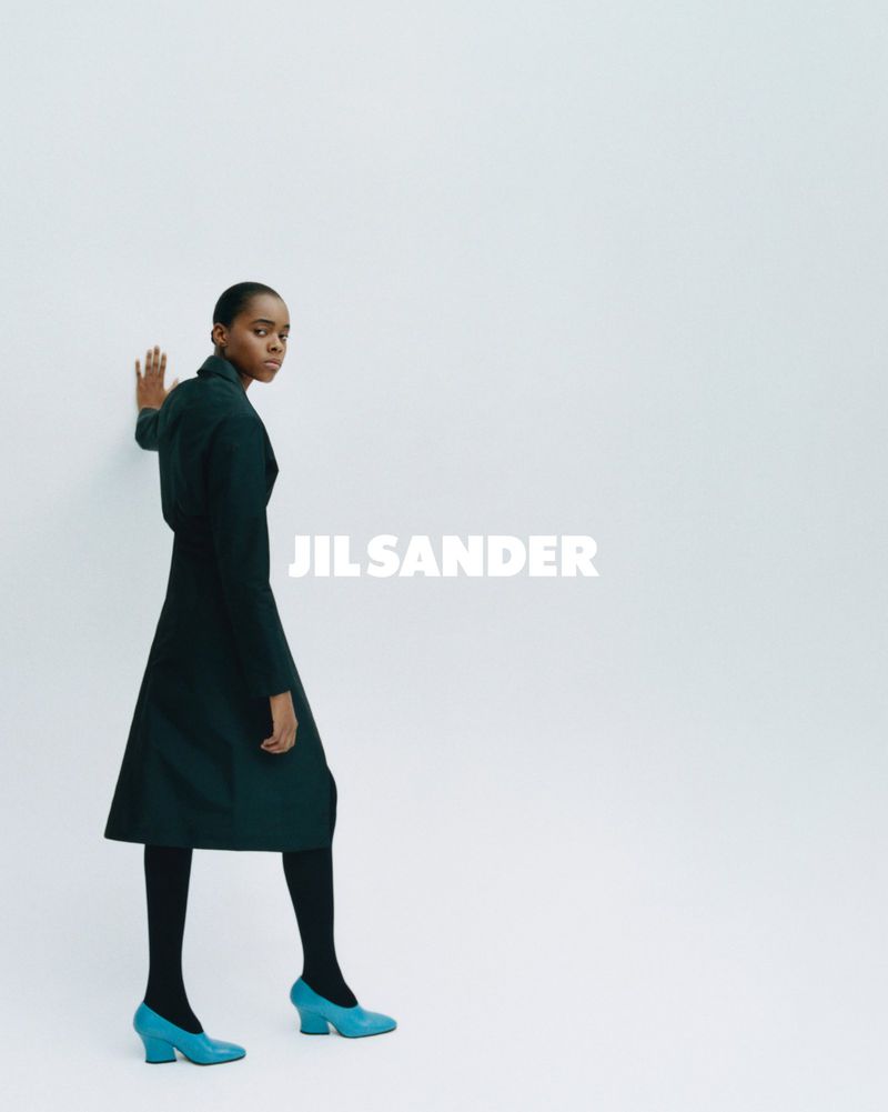 Model wearing dark green coat and bright blue heels against a white studio background with Jil Sander text overlay