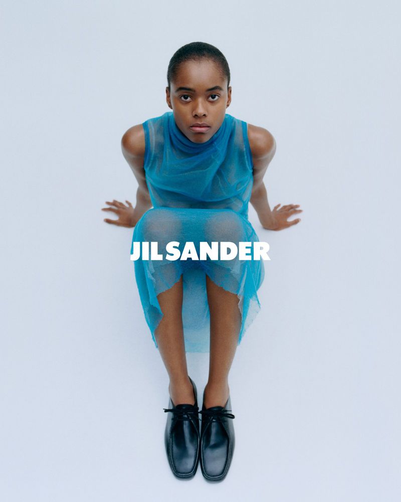 Fashion shot of a Black woman wearing a blue dress and black shoes, sitting against a white backdrop, with Jil Sander logo. 