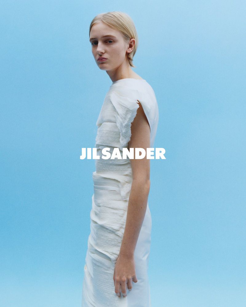 Fashion shot of a woman in a light-colored dress against a blue background, featuring the Jil Sander logo, showcasing a minimalist aesthetic.