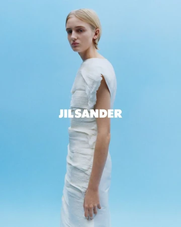 Fashion shot of a woman in a light-colored dress against a blue background, featuring the Jil Sander logo, showcasing a minimalist aesthetic.