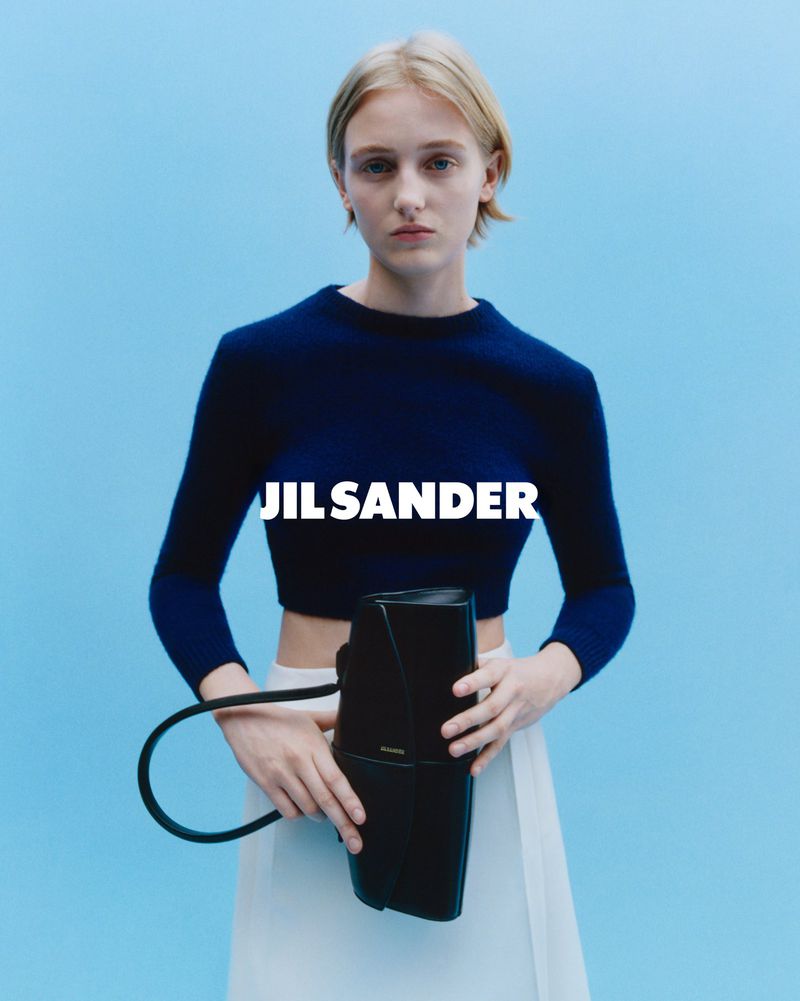 Model wearing dark blue top and white skirt holding black handbag against blue background