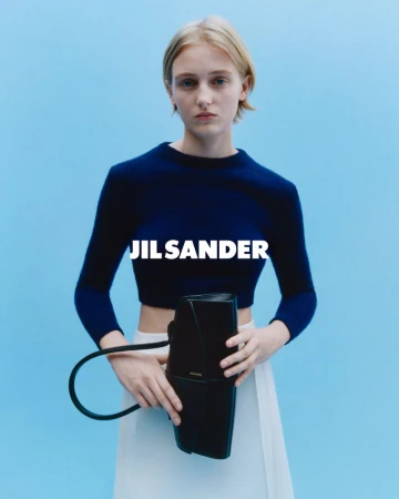 Model wearing dark blue top and white skirt holding black handbag against blue background