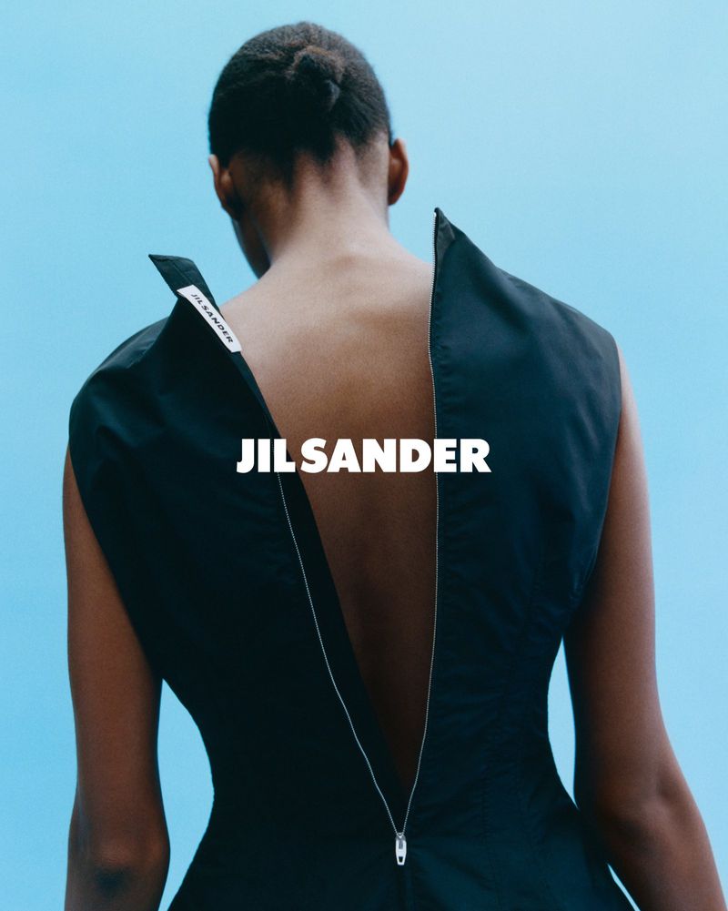 Back view of an adult woman wearing a sleeveless black dress with a zipper down the back against a light blue background. Fashion shoot.