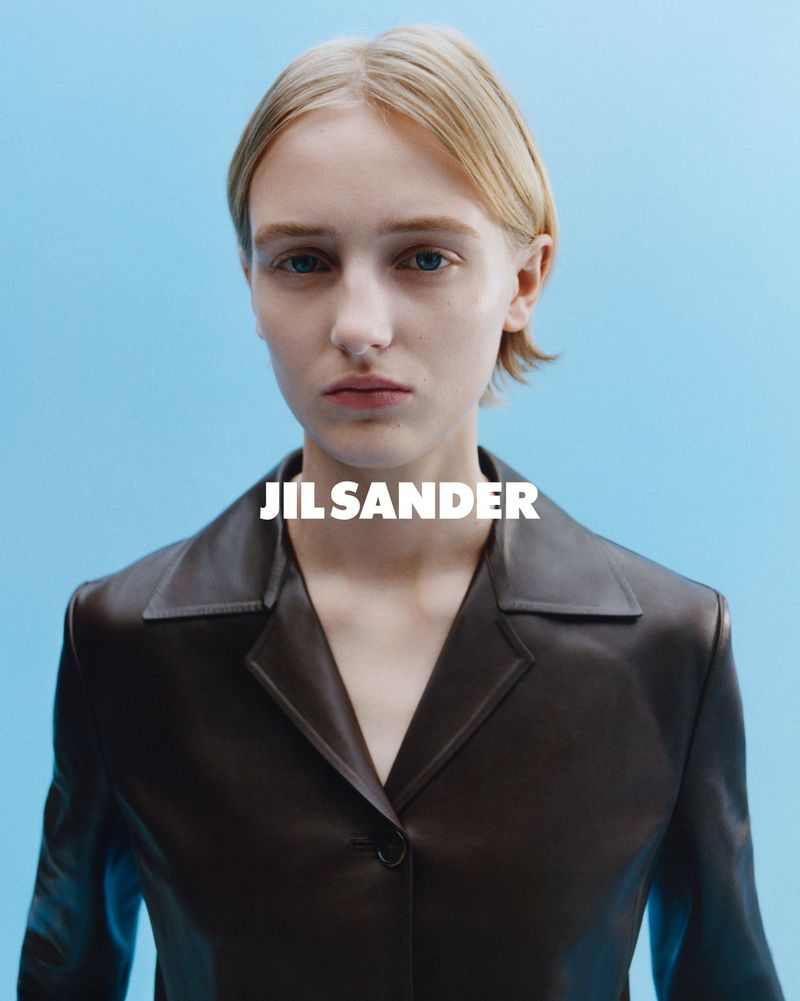 Close-up studio shot of a blonde fashion model in a brown leather jacket on a light blue backdrop with the brand name "JIL SANDER" visible.
