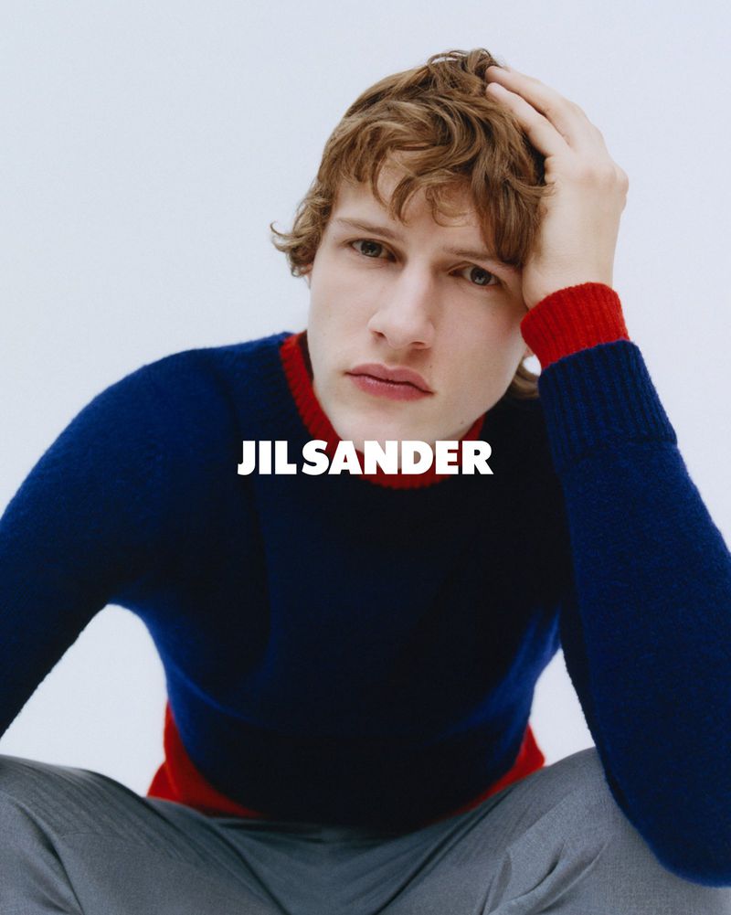 Young man in a relaxed pose, wearing a blue and red sweater with grey pants, set against a clean, bright backdrop for a fashion shot.