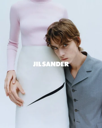 Fashion ad with a young adult man in grey blazer leaning against a woman in a pink turtleneck and white skirt, studio setting.