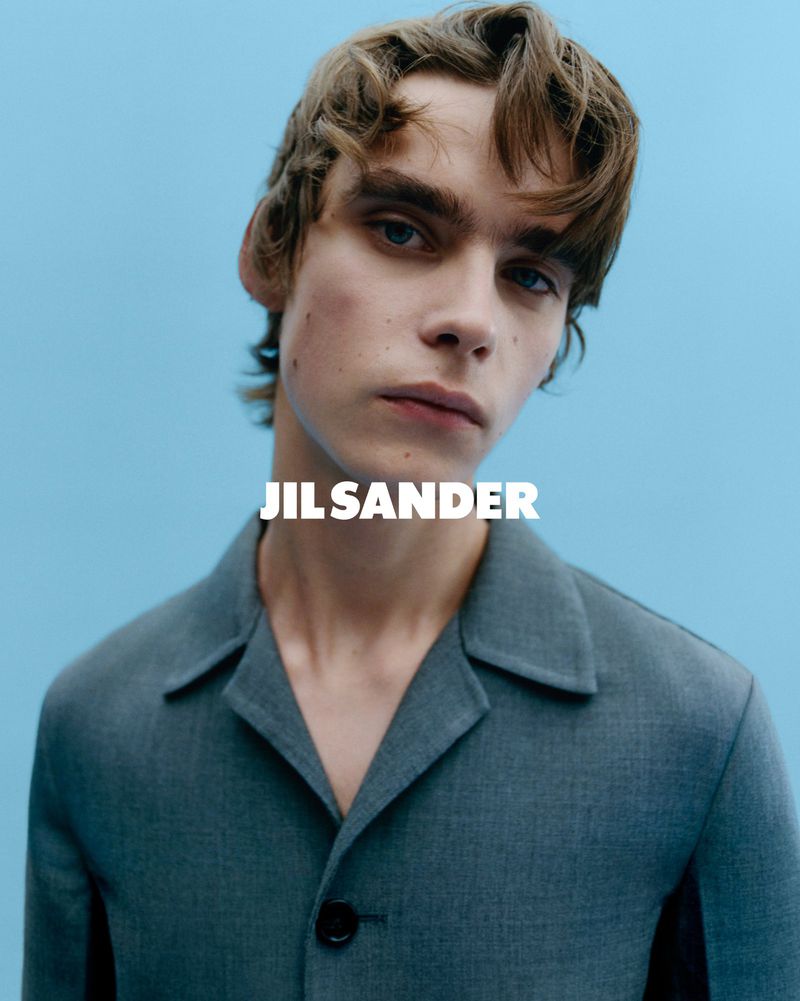 Portrait of a young man with wavy brown hair wearing a gray blazer against a blue background, featuring the Jil Sander logo.