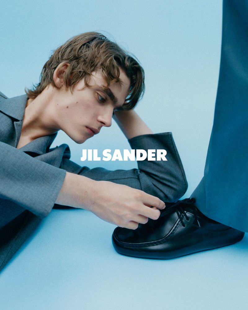 Fashion shot of a fair-skinned male model in a gray jacket looking at a black leather shoe against a blue background with "JIL SANDER" text.