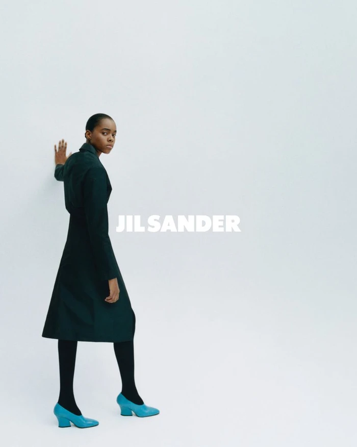 Model wearing dark green coat and bright blue heels against a white studio background with Jil Sander text overlay