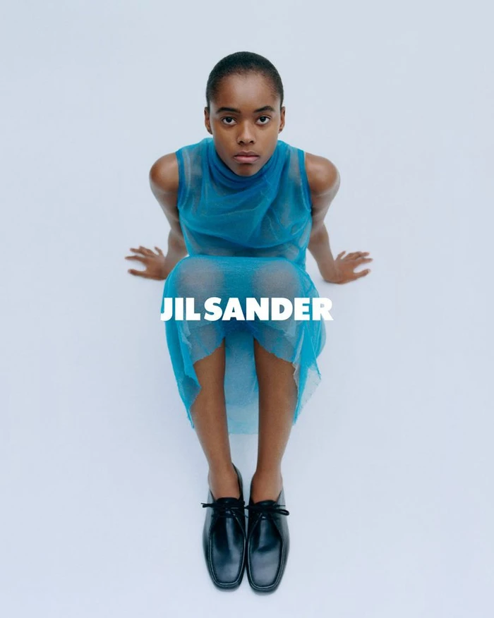 Model wearing a sheer blue sleeveless dress and black shoes seated against a plain white background for Jil Sander S/S 2026 campaign.