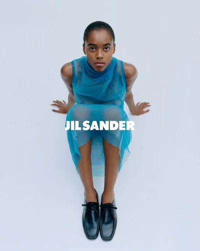 Model wearing a sheer blue sleeveless dress and black shoes seated against a plain white background for Jil Sander S/S 2026 campaign.