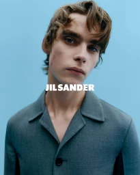 Close-up portrait of a young man wearing a grey collared button-up jacket against a solid light blue background.