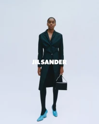Model standing against a white seamless background wearing a dark coat, tights, and bright blue shoes while holding a black handbag.