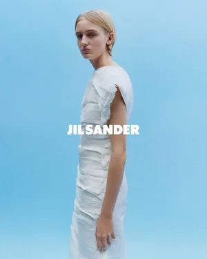 Model in a white sleeveless dress standing against a light blue seamless backdrop for Jil Sander S/S 2026 campaign.