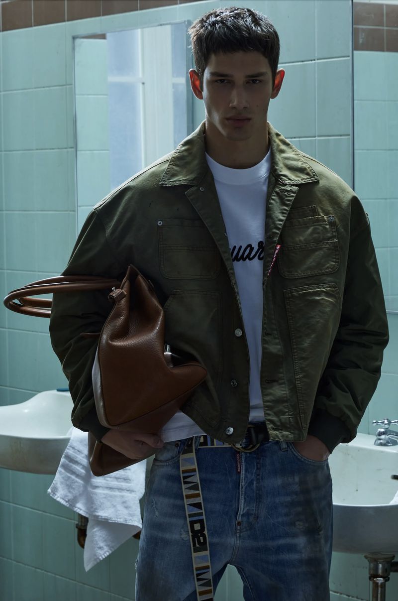 Fashionable man in bathroom wearing jacket, jeans, branded belt, holding tote bag; teal tile wall, utilitarian interior, medium shot.