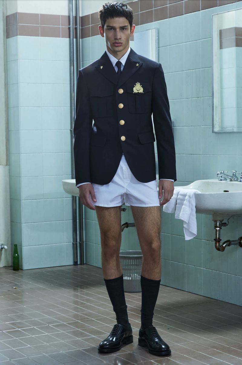 Man posing indoors wearing a black jacket with gold buttons and white shorts, showcasing a blend of formal and casual attire.