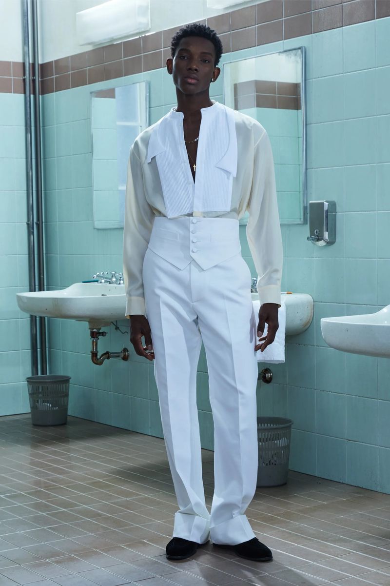 An adult male fashion model poses in a bathroom wearing a white shirt and pants with a vest, set against a backdrop of blue tiles and sinks.