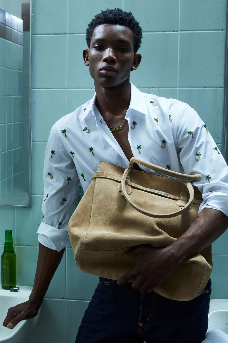 Portrait of Black male model in bathroom, holding large tan suede tote bag, wearing pineapple-print shirt and dark jeans, next to a beer bottle.