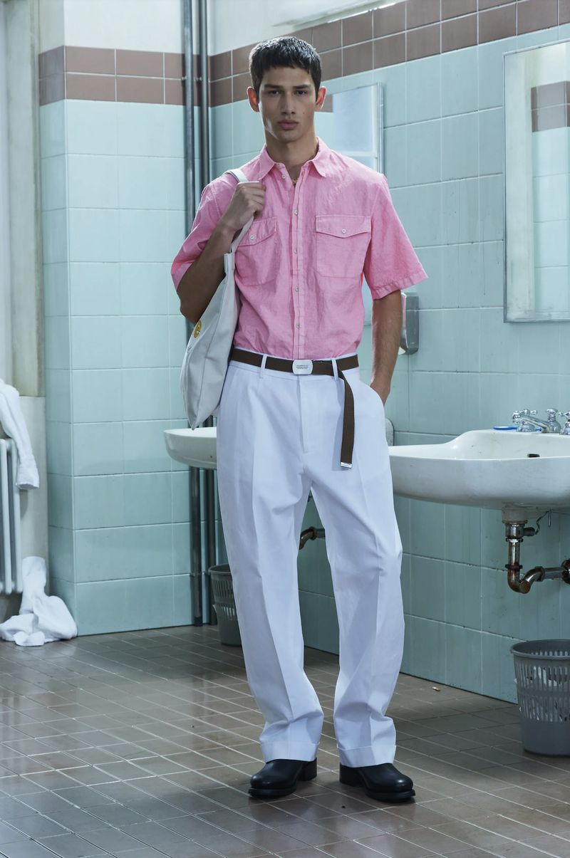 A young adult male is standing in a bathroom wearing a pink short sleeve shirt, white pants, and black shoes, carrying a white tote bag.