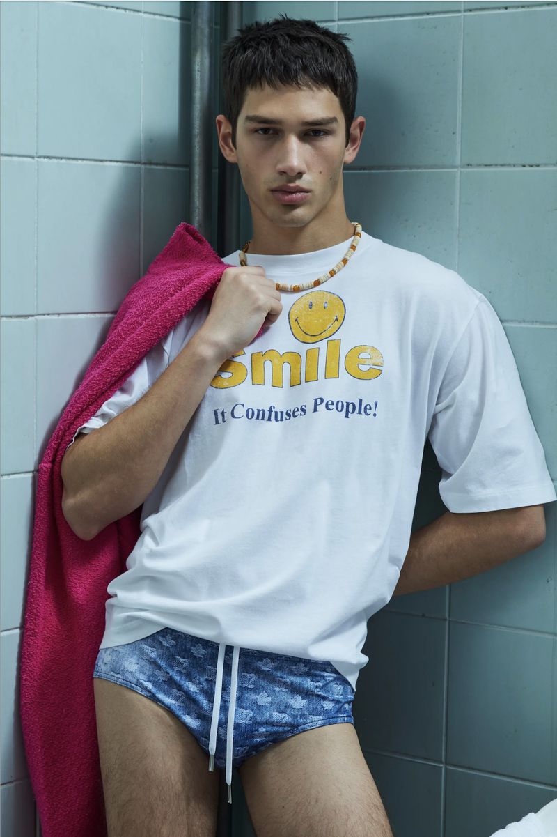 Young man poses against blue tile backdrop. He wears blue swim trunks and a white t-shirt with smile graphic. A pink towel is draped over his shoulder.