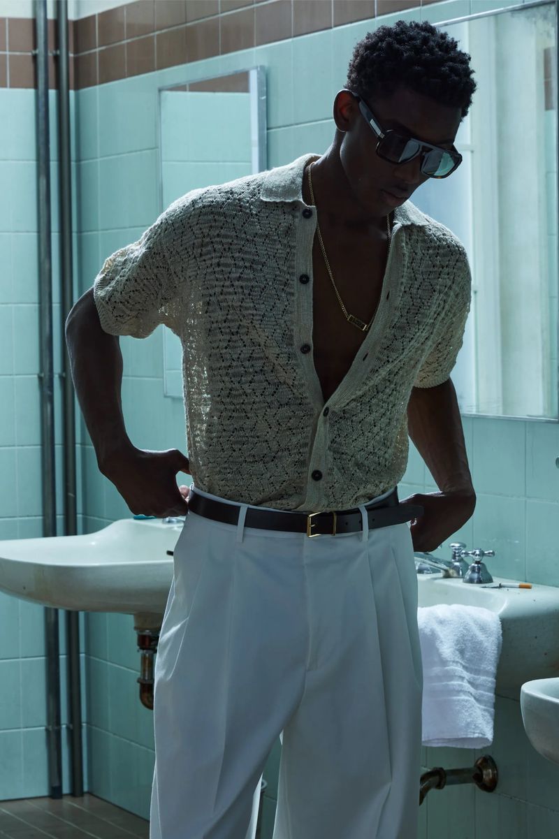 A stylish African-American man poses in a bathroom wearing a knit shirt, white pants, and sunglasses, showcasing fashion in an indoor setting.