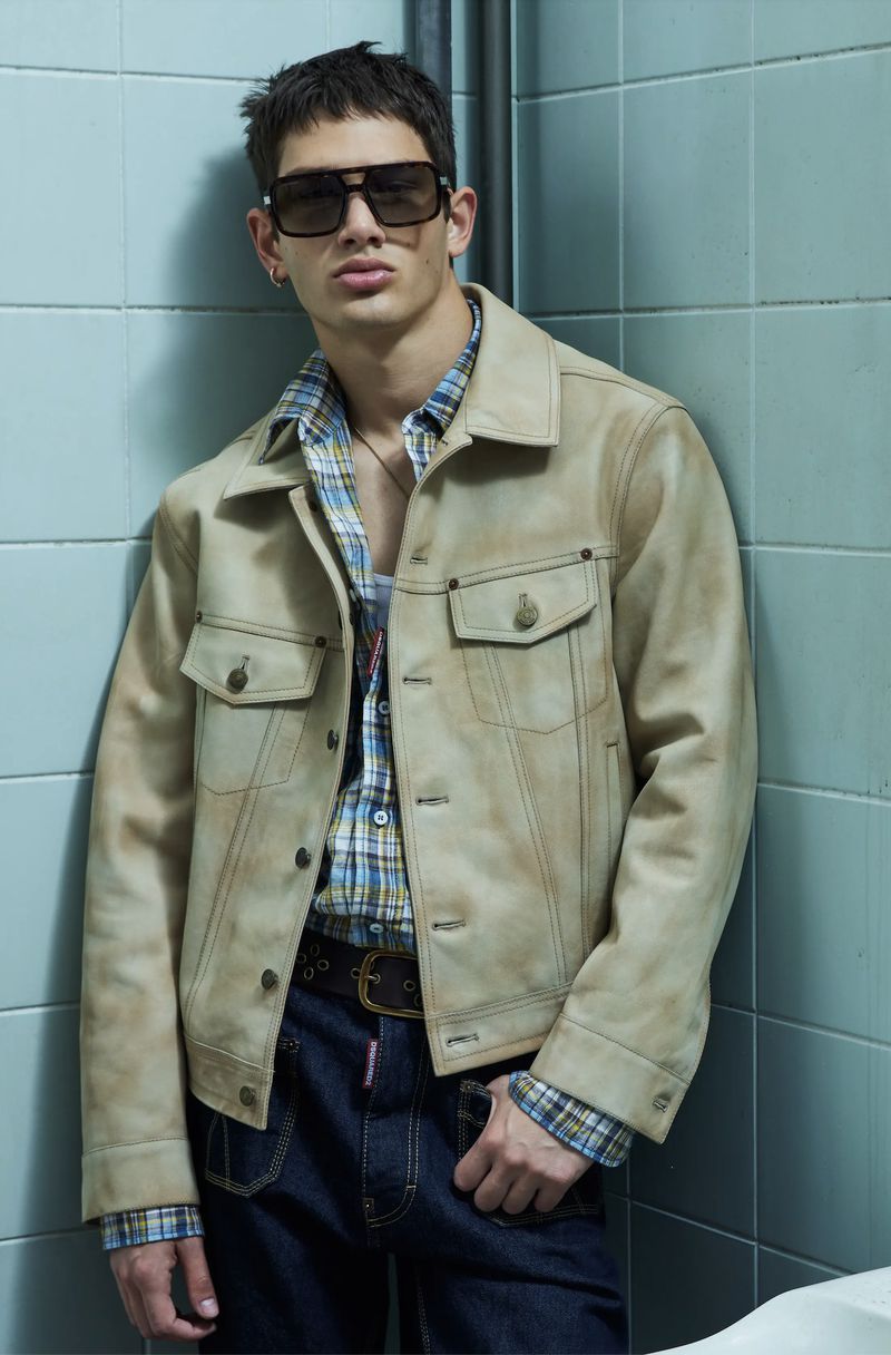 Fashionable adult man with sunglasses and a jacket poses against a blue tiled wall in a medium shot. He's wearing a plaid shirt and jeans.
