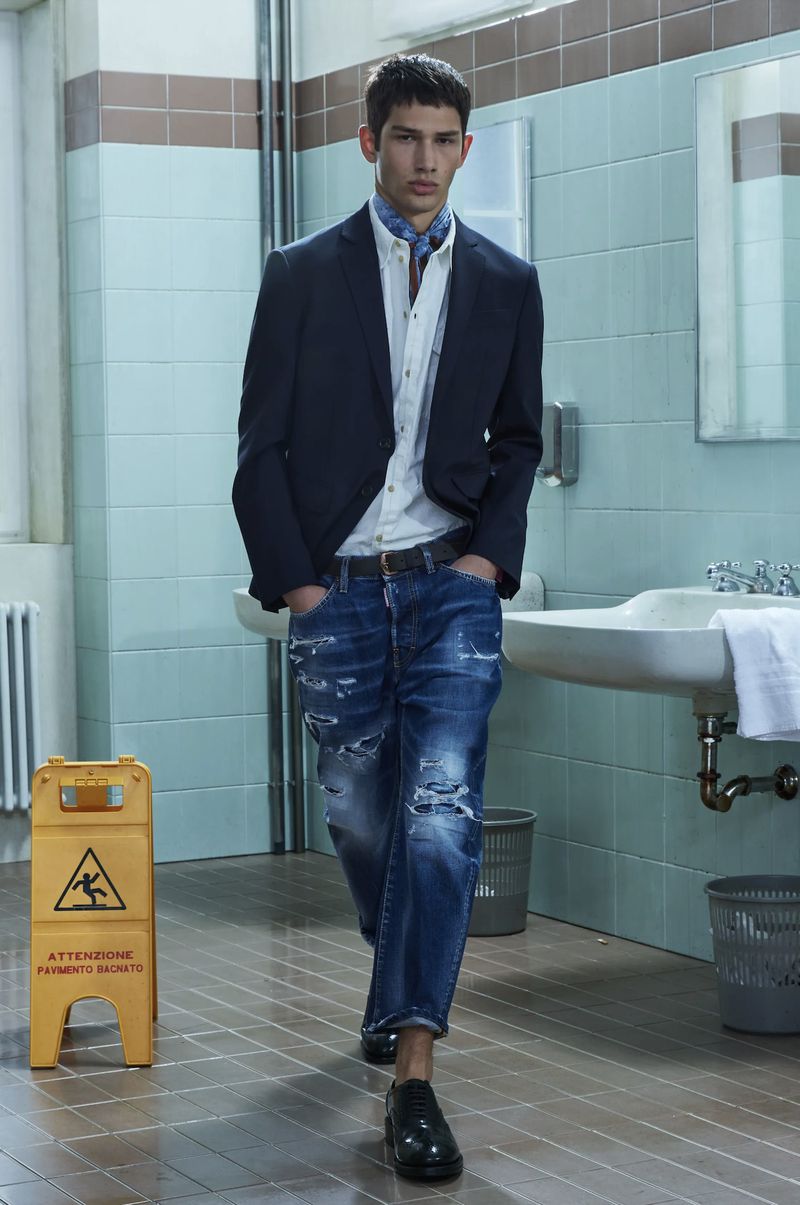 An adult male fashion model poses in a navy blazer, white button-down, distressed denim jeans, and black shoes in a tiled bathroom setting.