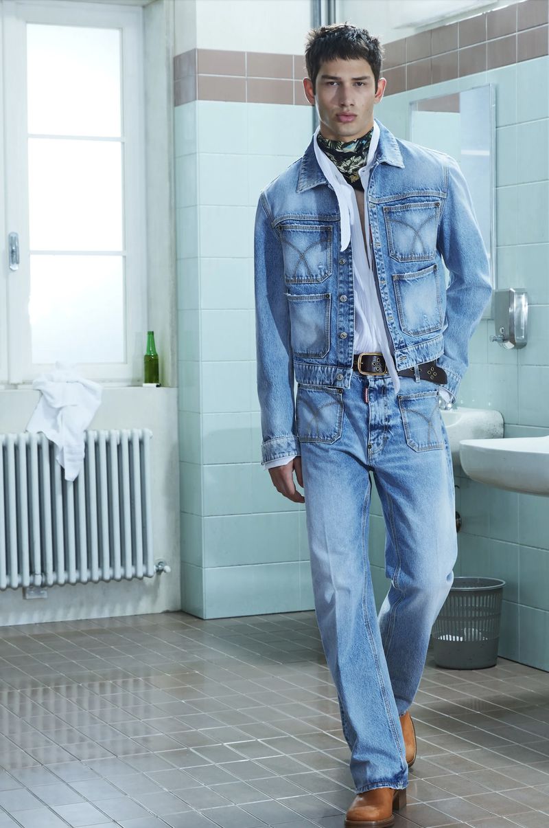 A stylish adult male stands indoors on tiled floor in a light blue denim jacket, jeans, a white shirt, and brown heeled boots.