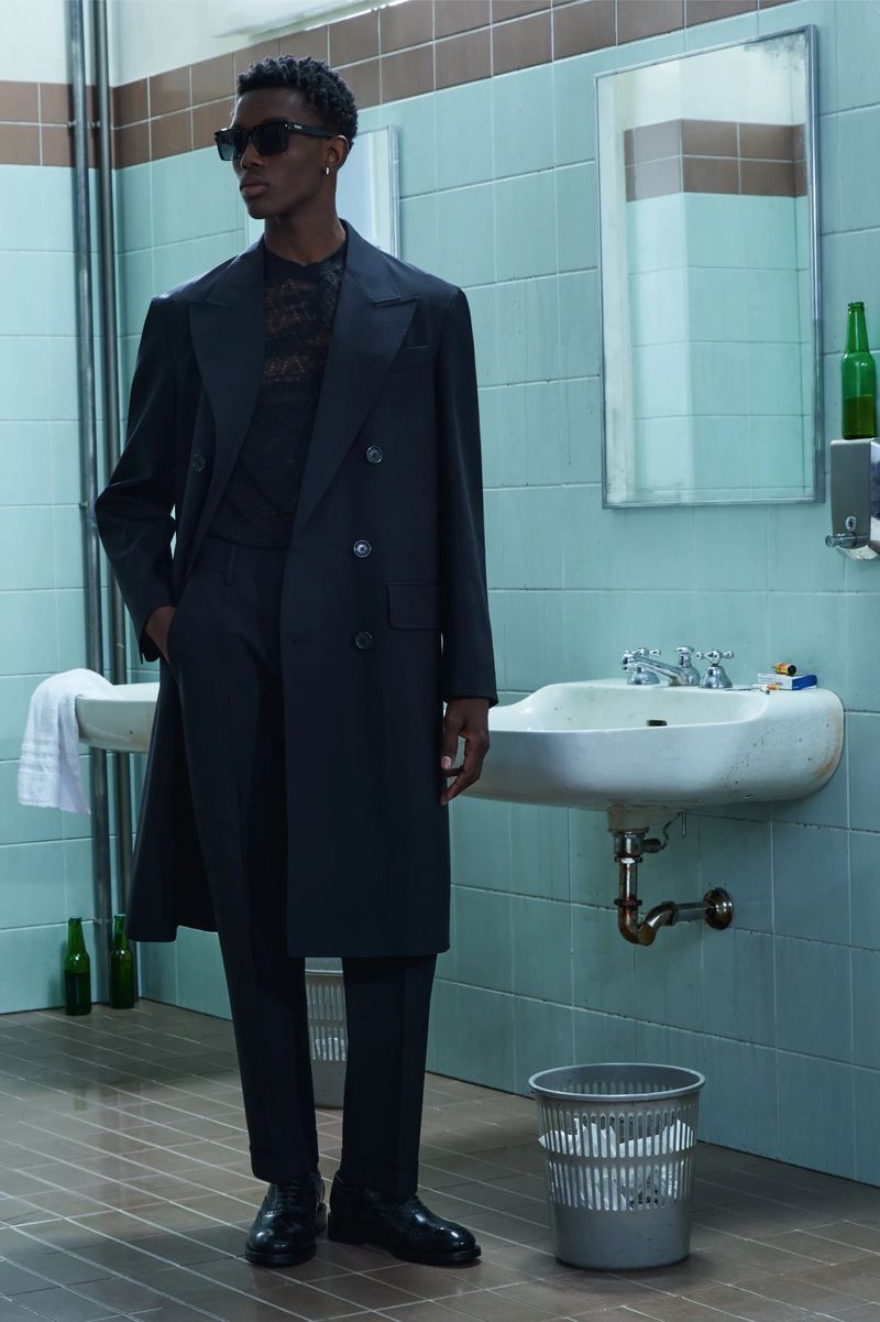 A slender Black male model strikes a pose indoors, wearing a dark suit and sunglasses in a grungy bathroom with blue tiles and green bottles.