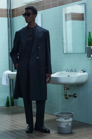 A slender Black male model strikes a pose indoors, wearing a dark suit and sunglasses in a grungy bathroom with blue tiles and green bottles.
