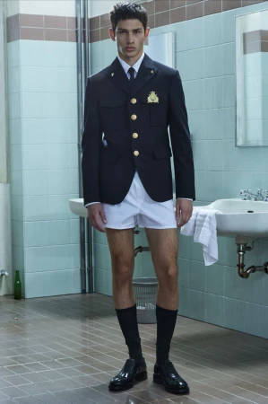 Model standing in a tiled restroom wearing a dark blazer, white shorts, black knee socks and dress shoes for Dsquared2.