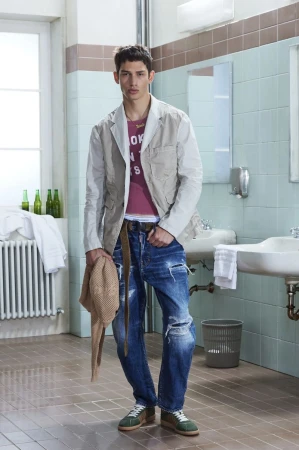 Model standing in a tiled restroom wearing a grey jacket, red t-shirt, and distressed denim jeans for Dsquared2 Resort 2026 Lookbook